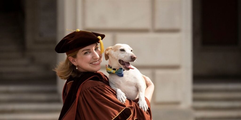 The image shows a person wearing academic regalia, including a dark brown velvet cap with a gold tassel and a matching gown. They are smiling and holding a small, white dog wearing a multicolored bow tie.