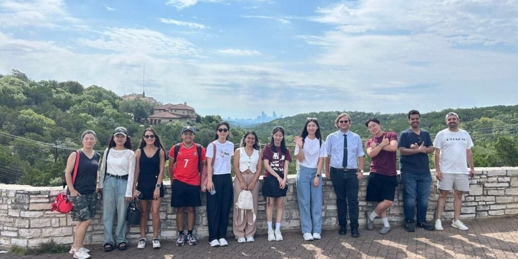 Group of twelve diverse students standing against a low stone wall backdropped by a lush green tree filled hills and a cloudy sky