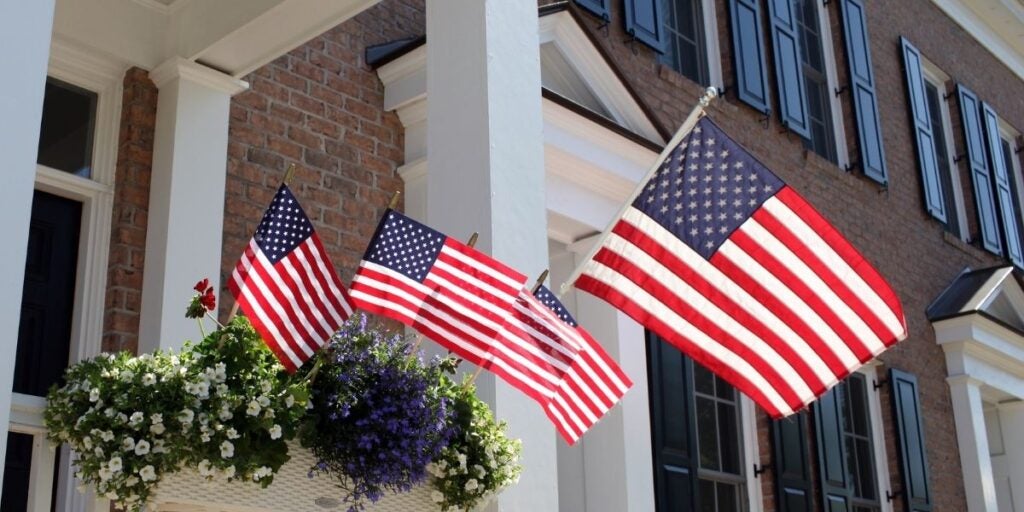 Three American flags are displayed outside a brick building with tall windows. A flower basket hangs nearby, adding vibrant green, white, and purple colors.